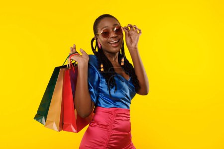 Téléchargez les photos : Shopping. Black Female Holding Shopper Bags Posing Wearing Sunglasses Smiling To Camera Standing Over Yellow Background, Studio Shot. Seasonal Summer Sales Advertisement - en image libre de droit