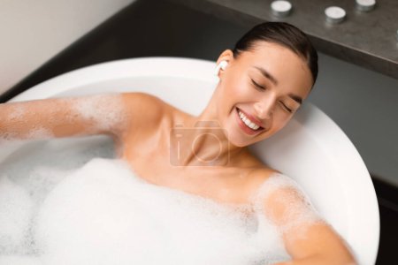 Photo for Relaxed Woman Listening To Music In Earbuds Lying In Bath With Foam In Bathroom Indoors, View From Above. Female With Eyes Closed Enjoying Favorite Song Online Bathing At Home. Relaxation Concept - Royalty Free Image