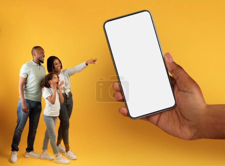 Photo for Excited Black Family Of Three Pointing At Big Blank Smartphone In Giant Female Hand While Walking Together Over Yellow Background, Happy Parents And Daughter Enjoying Online Offer, Mockup - Royalty Free Image