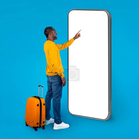 Photo for Smiling african american man in casual outwear with suitcase tourist touching with finger big smartphone white blank screen and smiling, black guy traveler check-in online, mockup, copy space - Royalty Free Image