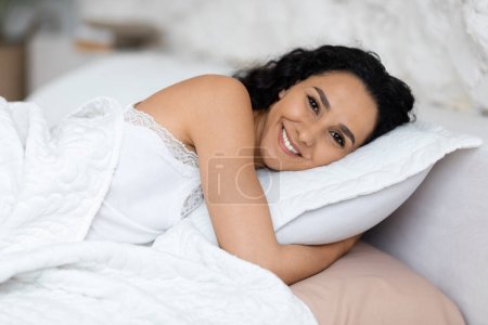 Téléchargez les photos : Glad femme caucasienne millénaire se réveiller et se couche sur un oreiller doux, profiter de bonne matinée, se reposer et se détendre dans l'intérieur minimaliste chambre blanche. Vitalité et doux rêves le week-end, temps libre, annonce et offre - en image libre de droit