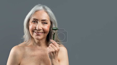 Téléchargez les photos : Cheerful pretty aged european lady with gray hair shows jade roller for face massage, isolated on gray background, studio, panorama. Beauty care, spa treatments, anti-wrinkle procedure, ad and offer - en image libre de droit