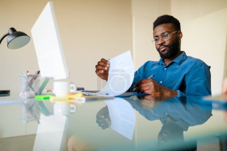 Téléchargez les photos : Homme d'affaires noir travaillant au bureau avec des documents et des ordinateurs, faisant de la paperasserie ou vérifiant des rapports financiers, assis sur le lieu de travail, espace libre - en image libre de droit