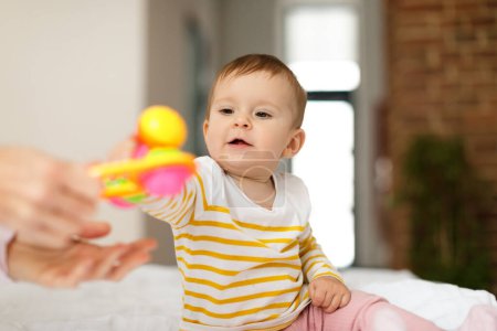Foto de Cute baby girl taking rattle toy from mother hands, sitting on bed, little child in bodysuit resting at home with mom, copy space. Child care concept - Imagen libre de derechos