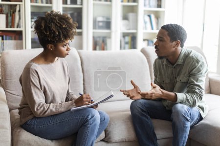 Photo for Young african american woman therapist taking notes on her upset patient at therapy session, frustrated black man having conversation with psychologist, sharing feelings, counselor office interior - Royalty Free Image