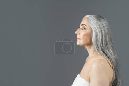 Téléchargez les photos : Serious retired european lady with gray hair look at copy space, isolated on gray background, studio, profile. Beauty and skin care, wrinkle treatment, anti-aging procedure, cosmetology, ad and offer - en image libre de droit