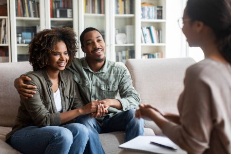 Photo for Happy beautiful young black couple in casual sitting on couch in cozy counselor office, embracing, holding hands and smiling after successful marital family therapy, copy space - Royalty Free Image
