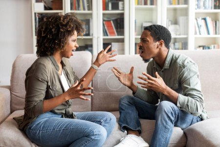 Foto de Furiosa pareja afroamericana discutiendo en el sofá en casa en la sala de estar, gritándose y haciendo gestos. Los esposos emocionales que visitan al psicólogo, que discuten - Imagen libre de derechos