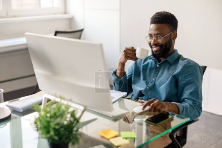 Foto de Sonriente hombre de negocios negro bebiendo café mientras trabaja en la computadora en el lugar de trabajo, mirando el monitor y sonriendo, leyendo el correo electrónico de negocios, charlando con los clientes en línea - Imagen libre de derechos
