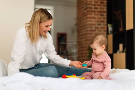 Foto de Developmental activities for babies. Mother playing with baby and holding toys, spending time with her cute infant child, sitting on bed at home, copy space. Mom and baby having good time together - Imagen libre de derechos