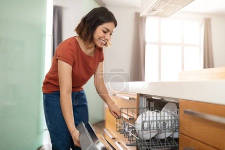 Photo for Housekeeping Concept. Young Smiling Arab Woman Opening Dishwasher Machine In Kitchen At Home, Happy Middle Eastern Female Unloading Clean Dishes And Cutlery, Doing Domestic Chores , Copy Space - Royalty Free Image