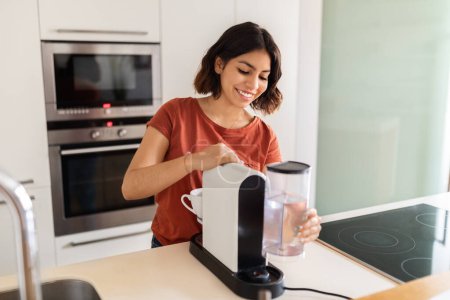 Téléchargez les photos : Entretien des appareils ménagers. Souriant jeune femme arabe remplissant la machine à café avec de l'eau dans la cuisine, Happy Millennial Middle Eastern Female Préparation de la boisson à la caféine à la maison, Espace de copie - en image libre de droit
