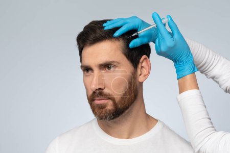 Foto de Middle aged man getting hair treatment at beauty salon, having mesotherapy session, standing on grey background. Female therapist hands in protective gloves making injection with syringe - Imagen libre de derechos