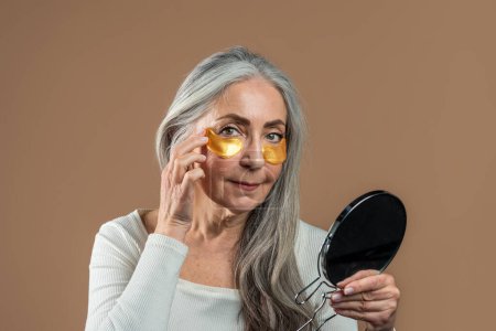 Foto de Glad mature european female with gray hair with golden patches under eyes enjoys spa treatments, looks in mirror isolated on brown background, studio. Anti-aging, anti-wrinkle procedure, beauty care - Imagen libre de derechos