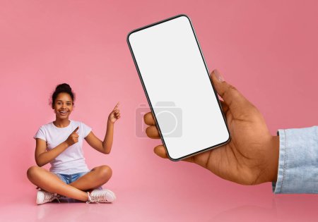 Photo for Nice Offer. Excited Black Teen Girl Pointing At Big Blank Smartphone In Huge Hand, Cheerful African American Female Child Showing Copy Space On Empty Cellphone, Sitting Over Pink Background, Mockup - Royalty Free Image
