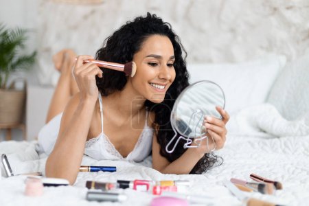 Photo for Glad millennial caucasian brunette lady with long hair lies on soft bed applies powder with brush on face, looks in mirror in minimalist bedroom interior. Nude makeup and routine at home, good morning - Royalty Free Image