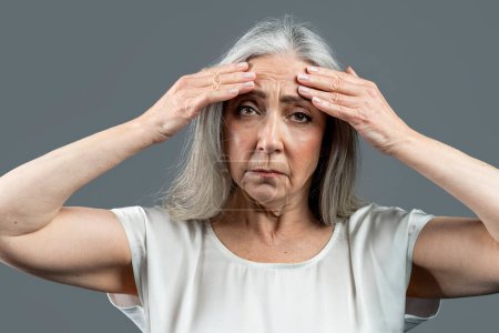 Foto de Sad old european lady with gray hair touches her face, wrinkles with hands, isolated on gray background, studio, copy space. Problems with beauty care, spa treatments, age-related changes at menopause - Imagen libre de derechos