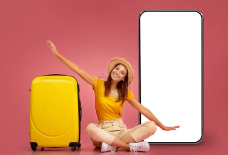 Téléchargez les photos : Jolie jeune femme excitée en chapeau et vêtements décontractés confortables assis sur le sol par une valise jaune et un grand téléphone portable avec écran blanc, imitant l'avion, fond de studio rose, maquette - en image libre de droit