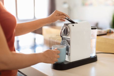 Téléchargez les photos : Jeune femme utilisant une machine à café tout en faisant une tasse de café à la maison, femme méconnaissable préparant une boisson à la caféine avec un appareil moderne dans la cuisine, appréciant le début de la journée, image recadrée - en image libre de droit