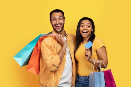 Photo for Cheerful shocked young african american male and lady hold many packages of purchases, show credit card, isolated on yellow background, studio. Cashback, shopping together, huge sale and finance - Royalty Free Image