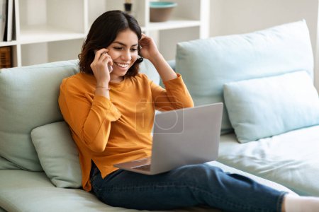 Téléchargez les photos : Jeune femme arabe heureuse parlant sur son téléphone portable et faisant du shopping en ligne sur son ordinateur portable à la maison, souriante femme du Moyen-Orient faisant des achats sur Internet via ordinateur et bénéficiant d'un appel téléphonique, espace libre - en image libre de droit