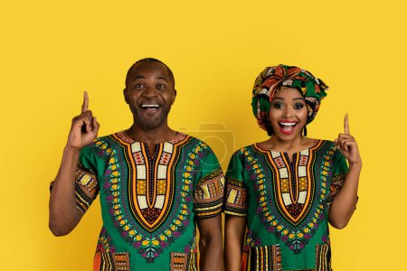 Foto de Amar atractivo adulto negro hombre y mujer en brillante nacional africano ropa posando en amarillo fondo del estudio, apuntando hacia arriba en el espacio de copia de arriba, mostrando agradable oferta en línea - Imagen libre de derechos