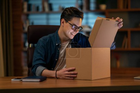 Photo for Smiling man sitting at workplace home office desk wearing glasses unpacking opening cardboard box long awaited parcel with goods bought on-line. Satisfied client of shipping delivery service concept - Royalty Free Image