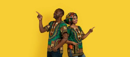 Photo for Positive happy cheerful black middle aged man and young woman in national african clothing standing back to back, pointing at copy space over yellow studio background, web-banner - Royalty Free Image