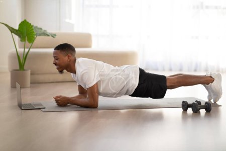 Foto de Side view of motivated smiling young black guy in sportswear planking on yoga mat at home, watching fitness video on Internet. African american man exercising after waking up. Healthy lifestyle - Imagen libre de derechos