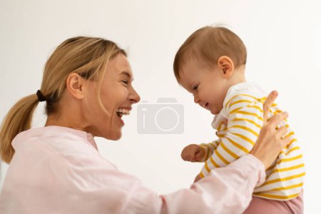 Téléchargez les photos : L'amour maternel. Mère caucasienne heureuse tenant mignonne petite fille sur les mains et la couette, regardant bébé fille et souriant sur fond de mur blanc, vue de côté - en image libre de droit