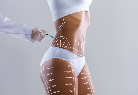 Foto de Millennial slim black lady in white shorts with abstract lines and arrows gets injection from doctor isolated on gray wall background, studio. Beauty care, body shaping, medical care and buttocks - Imagen libre de derechos