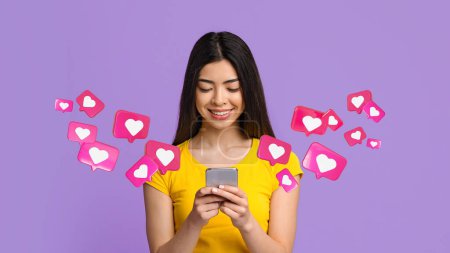 Foto de Alegre adolescente japonesa estudiante tiene charla romántica con los corazones en el teléfono, disfruta de mensajes, mensajes de texto y redes sociales sobre fondo púrpura, estudio, panorama. Aplicación para las relaciones y el amor, citas - Imagen libre de derechos