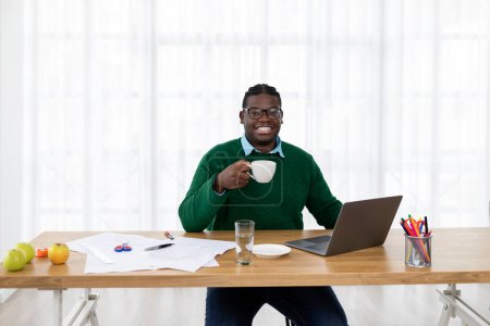 Téléchargez les photos : Heureux homme d'affaires afro-américain à l'ordinateur portable posant dans son bureau, assis à table et tenant tasse de café. Designer masculin travaillant sur l'ordinateur au travail. Concept de travail indépendant et en ligne - en image libre de droit