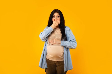 Téléchargez les photos : Portrait of young pregnant lady suffering nausea while standing over yellow studio background, expectant woman covering mouth with hand, feeling unwell, free space - en image libre de droit