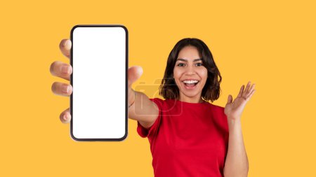 Photo for Emotional pretty young arab woman in red t-shirt showing brand new cell phone with white empty screen and gesturing over yellow studio background, online offer, mockup, panorama - Royalty Free Image