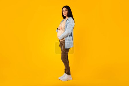 Foto de Mujer embarazada joven tocando el vientre mientras posando sobre fondo de estudio amarillo, tiro de cuerpo entero, espacio libre. Señora expectante abrazando barriga y sonriendo a la cámara - Imagen libre de derechos