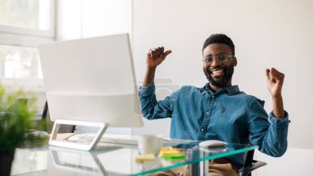 Photo for Excited black businessman gesturing yes in front of computer, celebrating success, sitting at workplace in office, panorama. Male entrepreneur making great deal, signing online contract - Royalty Free Image