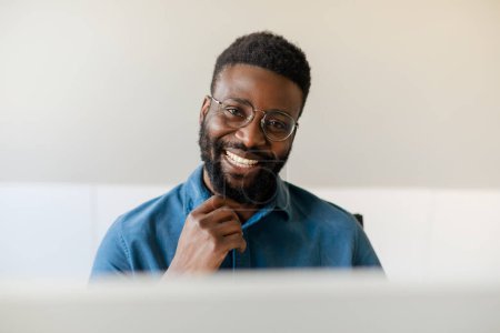 Téléchargez les photos : Le sourire du succès. Portrait d'un homme d'affaires afro-américain heureux travaillant dans son bureau, assis devant l'ordinateur et souriant à la caméra - en image libre de droit
