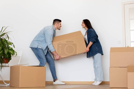 Téléchargez les photos : Happy Young Couple Carrying Big Cardboard Box With Bellongings At Home, Cheerful Millennial Spouses Moving To Their New House Together, Celebrating Relocation To Own Flat, Copy Space - en image libre de droit