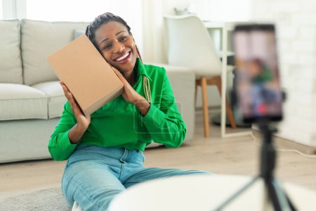 Zufriedene schwarze Frau umarmt Pappschachtel und dreht Videoblog auf Handy im Wohnzimmer Interieur. Arbeit zu Hause, moderne Technik und Auspacken während der Covid-19-Pandemie