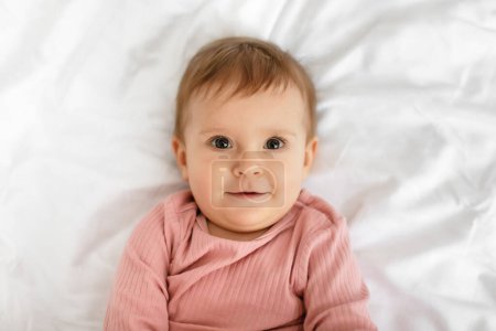Photo for Adorable caucasian baby in bodysuit lying on bed at home, resting on white bedsheets in bedroom, closeup portrait, free space, top view - Royalty Free Image