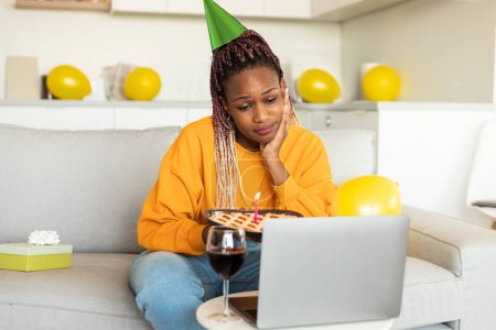 Foto de Triste mujer negra soltera en sombrero de fiesta celebrando cumpleaños en línea en cuarentena, utilizando el ordenador portátil para videollamada, sosteniendo pastel con vela, sentado en el sofá - Imagen libre de derechos