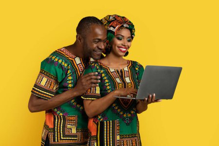 Photo for Beautiful cheerful smiling black couple wearing traditional african costumes using modern laptop together, man and woman posing on yellow studio background, surfing on Internet, copy space - Royalty Free Image