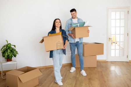 Foto de Happy young couple holding carton boxes and walking in their new apartment, cheerful millennial spouses carrying packages with belongings, celebrating relocation to own home, copy space - Imagen libre de derechos