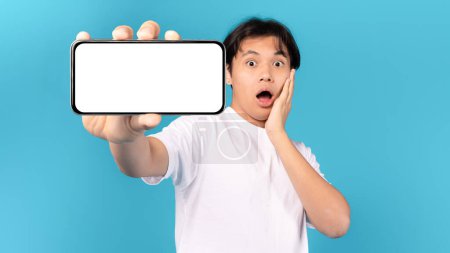 Photo for Shocked Japanese Teen Boy Showing Smartphone Blank Screen Touching Face Posing Standing Over Blue Studio Background. Omg, Great Mobile Offer And Application Ad Concept. Panorama, Mockup - Royalty Free Image