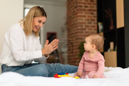 Photo for Happy mother smiling and playing clap hand with her adorable little daughter, sitting on bed at home. Mom and baby spending time together - Royalty Free Image