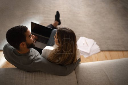 Photo for Young arabic male hug and look at european woman typing on computer with blank screen in living room interior with documents and charts. Business, work remotely together, love, relationship at home - Royalty Free Image