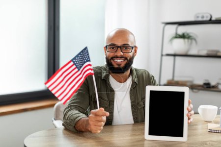 Foto de Happy latin man showing flag of the US and digital tablet with black blank screen, recommending educational mobile app or new website, sitting at desk, mockup - Imagen libre de derechos