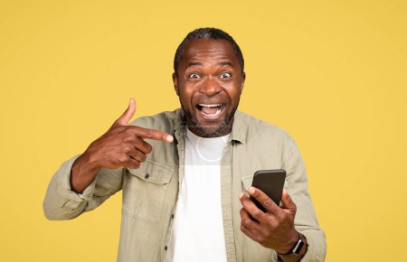 Photo for Glad excited shocked adult black guy in casual with open mouth pointing finger at smartphone, recommends app, isolated on yellow background, studio. Win and success, advice for blog, ad and offer - Royalty Free Image