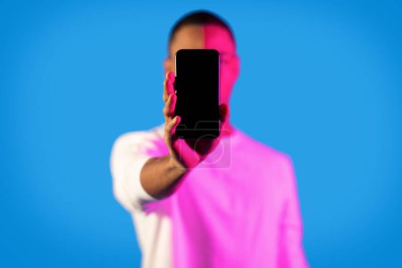 Téléchargez les photos : Unrecognizable black man in white longsleeve showing brand new smartphone with black empty screen at camera in neon light, african guy recommending online offer, blue background, mockup, copy space - en image libre de droit